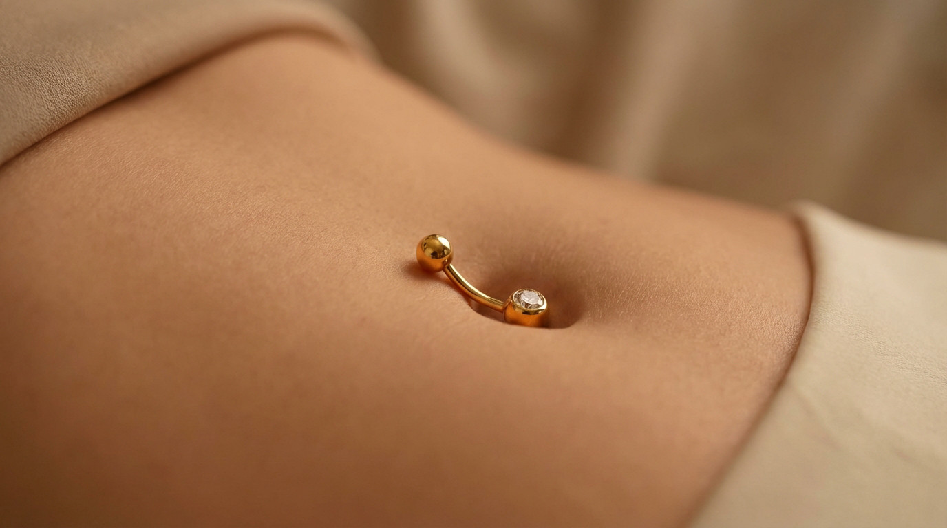 Close-up of an 18k gold navel piercing featuring a sparkling clear gemstone on smooth skin, with soft, warm lighting.